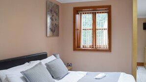 A neatly made bed with a headboard, two pillows, and a blanket. There is a window with wooden blinds, a painting on the wall, and a nightstand with a lamp. - George's Gaff - E4432 (Near Skegness)