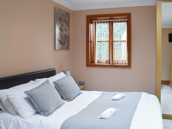 A neatly made bed with a headboard, two pillows, and a blanket. There is a window with wooden blinds, a painting on the wall, and a nightstand with a lamp.