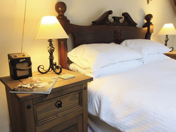 Een net opgemaakt bed met witte lakens, een houten nachtkastje met een lamp, een radio en een tijdschrift.
