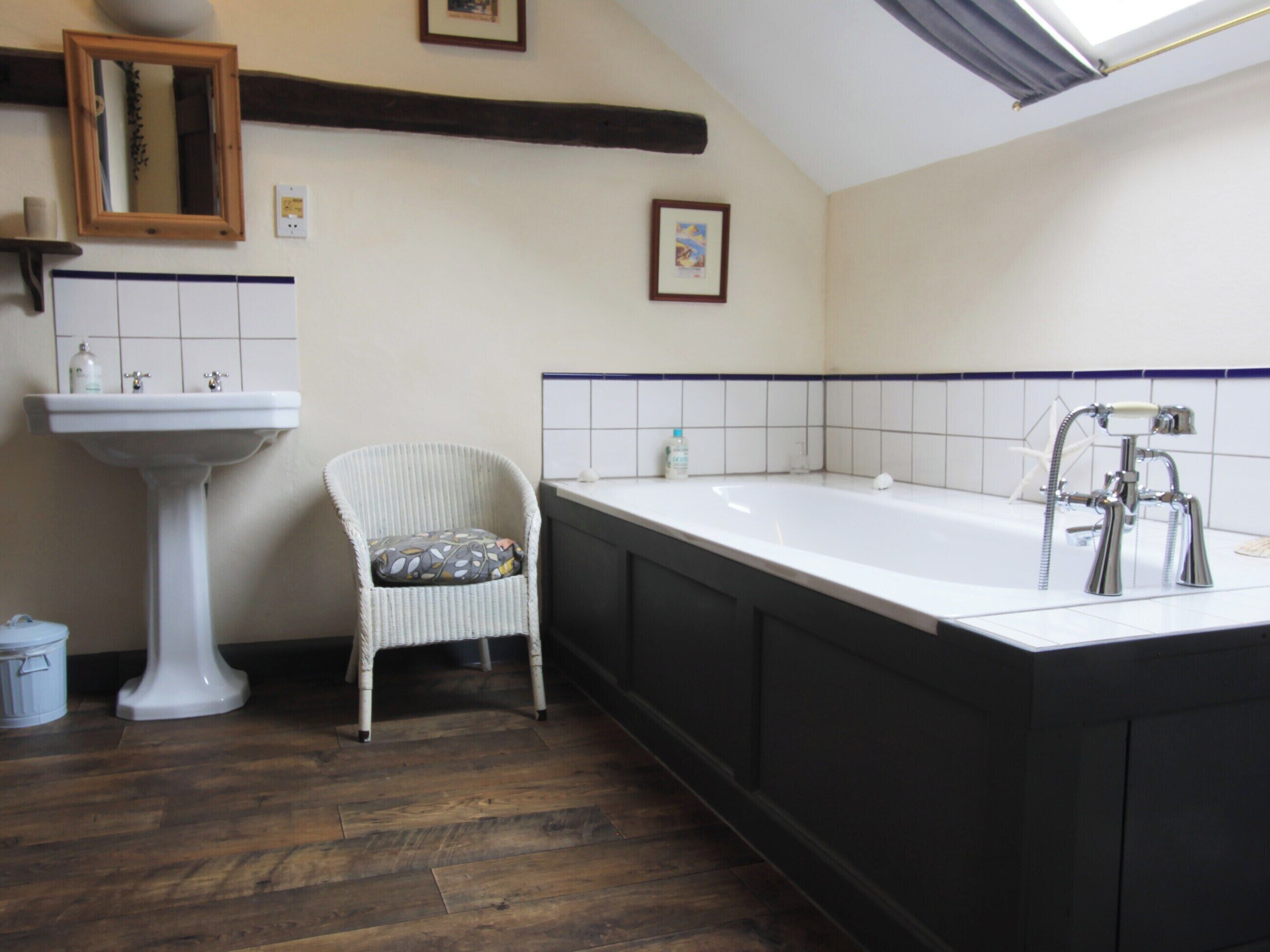 Un baño con lavamanos pedestal blanco, una silla de mimbre con cojín, una bañera grande y suelo de madera oscura.