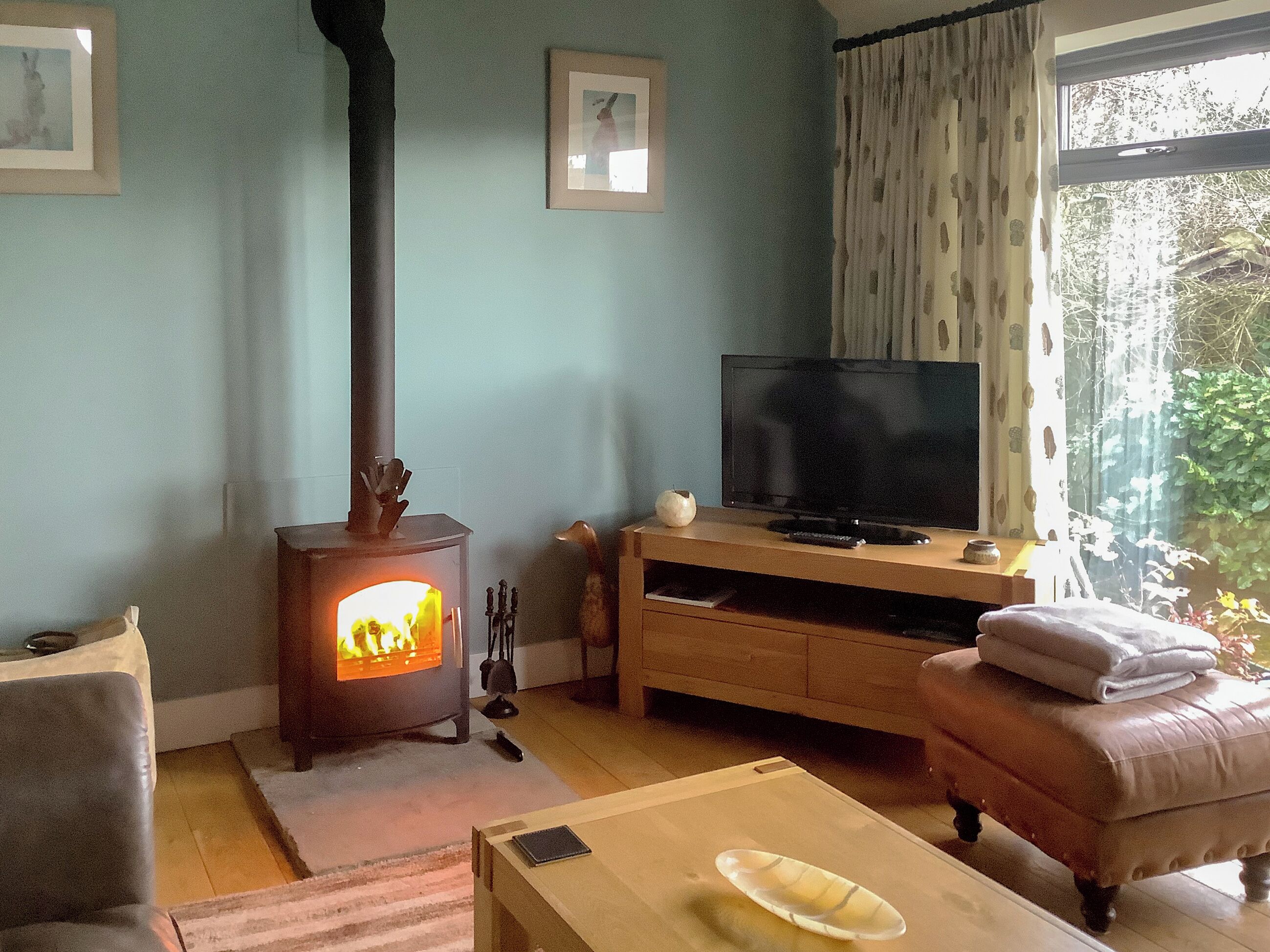 A living room with a wood-burning stove, a television on a wooden stand, a cushioned bench, and a window with curtains.