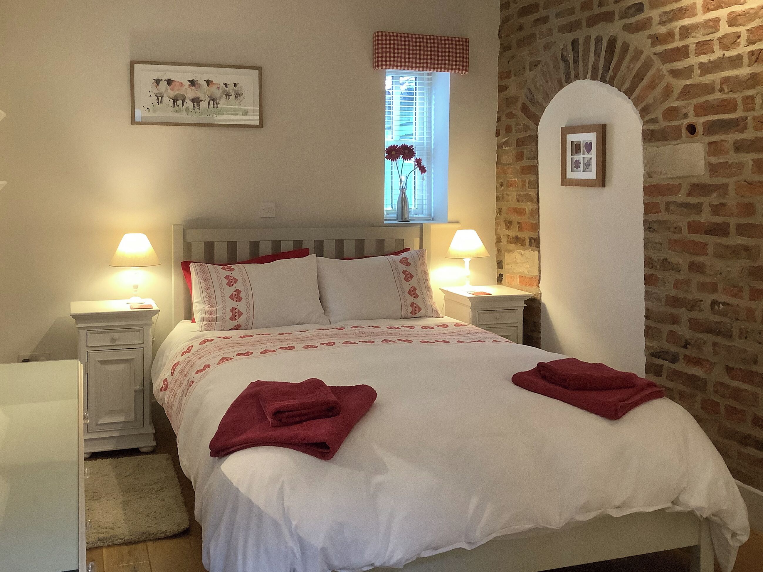 A neatly made bed with a white and red patterned bedspread, two red towels, and two bedside lamps.