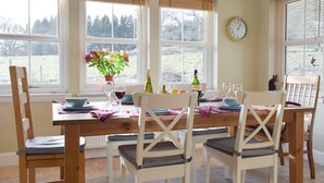 Un coin repas avec une table en bois dressée pour quatre personnes, des chaises blanches avec des coussins à carreaux, un vase rempli de fleurs et une horloge au mur.