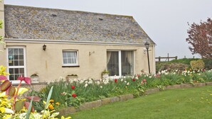 A house with a sloped roof, a front door, and a window. There is a garden with various plants and flowers. - 2 bedroom accommodation in Cosheston near Pembroke (Cosheston near Pembroke)