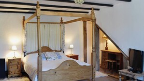 Un lit à baldaquin en bois, agrémenté de colonnes et d’un ciel de lit, flanqué de tables de chevet avec des lampes, dans une pièce dont les poutres apparentes sont en bois et qui est ornée d’un lustre.