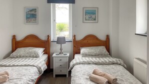 A room with two beds, a nightstand, a lamp, and two framed pictures on the walls. - 2 bedroom accommodation in St Austell (St Austell)