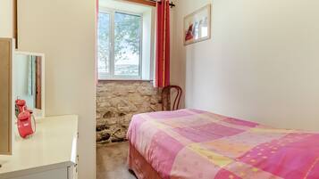 A bedroom with a bed, a dresser, a window with curtains, a stone wall, and a painting on the wall.