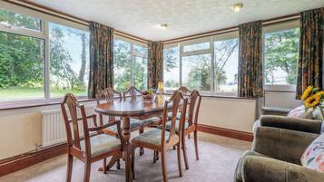 A dining area with a round table, wooden chairs, and a view of greenery through large windows.