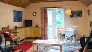 Ett mysigt vardagsrum med ett träpanelat tak, en tv på ett träskåp, en matplats med ett bord och stolar samt ett stort fönster med gardiner.