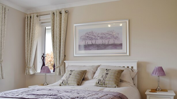 Une chambre à coucher avec un lit, deux tables de chevet, une fenêtre avec des rideaux, un tableau encadré au mur et une lampe de chevet.