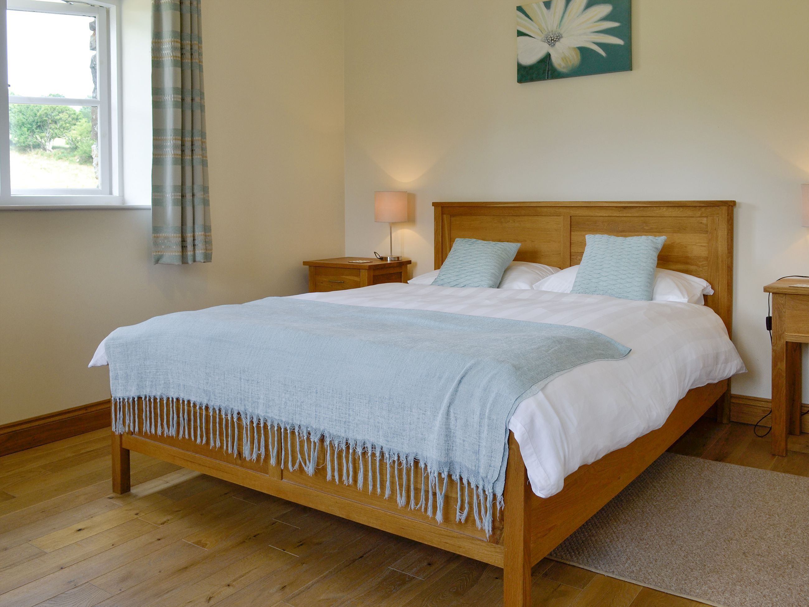 A bedroom with a wooden bed, a bedside table with a lamp, a window with curtains, and a wall painting.