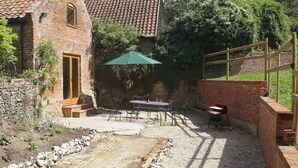 A rustic brick building with a tiled roof, a patio with a table and chairs, and a green umbrella providing shade. - The Street Farm Barn (Costessey, Norwich)