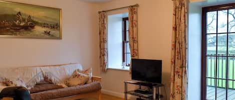 Una sala de estar con un sofá, un televisor sobre un mueble bajo, un cuadro en la pared y vistas a través de una ventana.