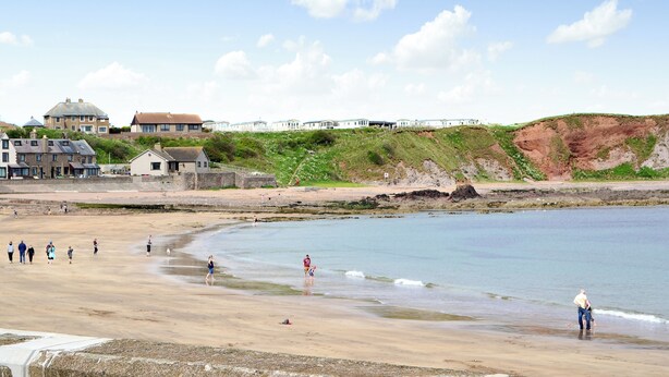 Ein Küstenstrand mit Menschen, die am Wasser entlanggehen und spielen, ein sandiger Uferstreifen und Gebäude im Hintergrund.
