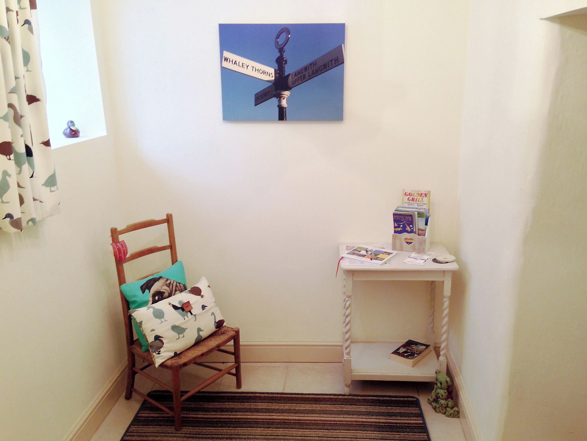 Una habitación de esquina con una silla de madera, una mesita con libros, una alfombra y un cartel en la pared.