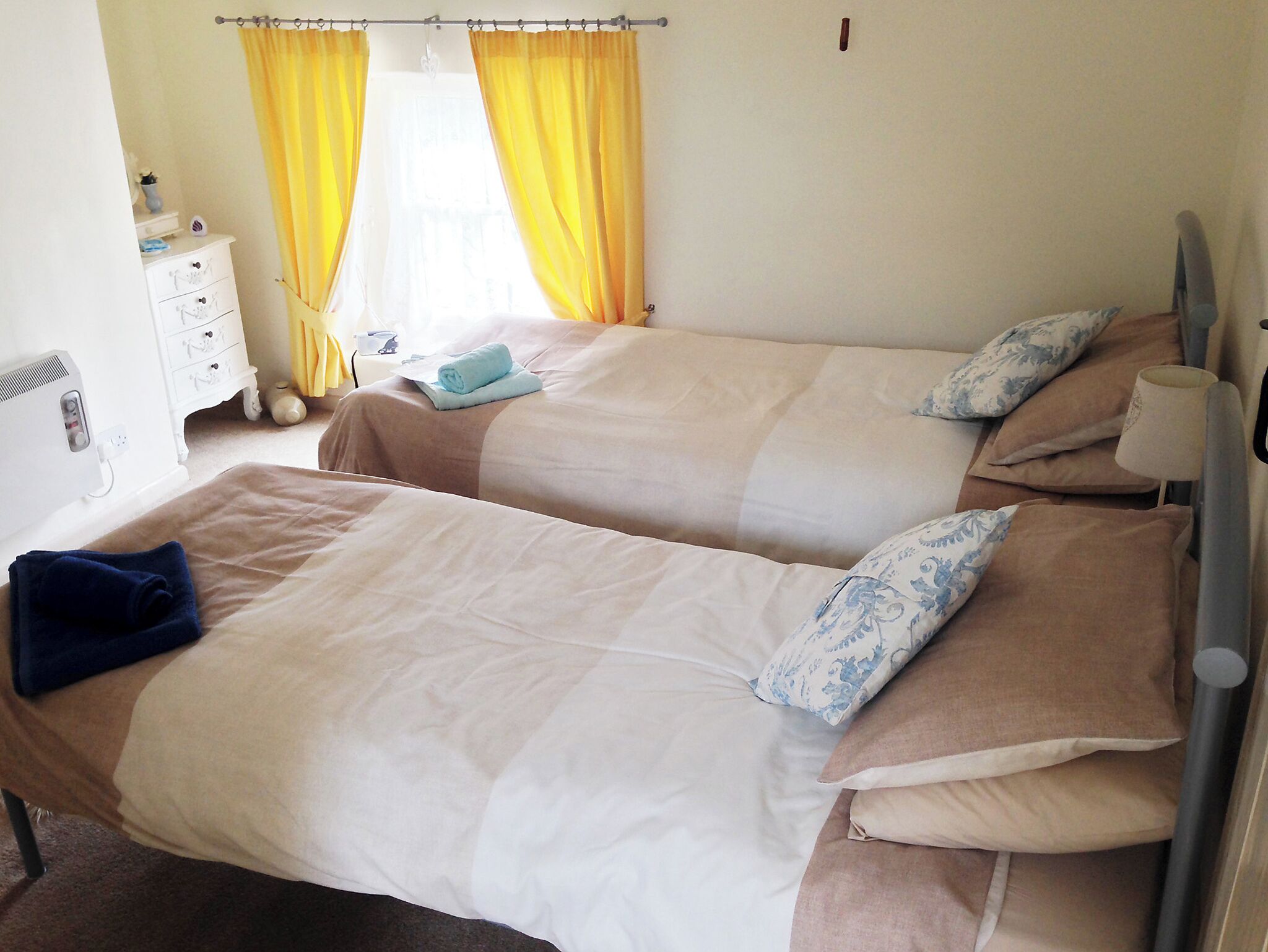 Une chambre d’hôtel avec deux lits, une table de chevet, une lampe et une fenêtre avec des rideaux jaunes.