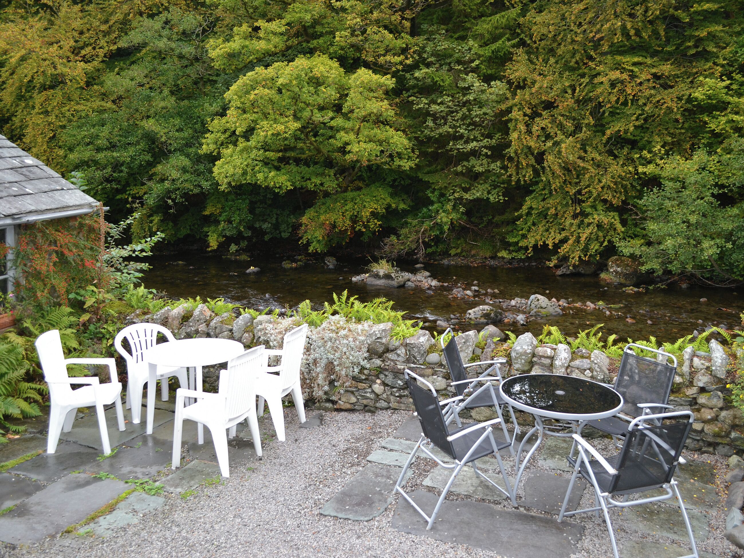 Un espace extérieur aménagé avec des chaises en plastique blanc et une table en métal, entouré d’un mur de pierre et offrant une vue sur une rivière et une forêt.