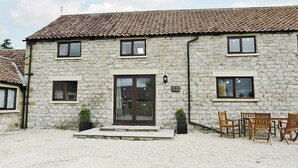 A stone cottage with a tiled roof, a front door, and a sign reading "Ash Tree Cottage." There are outdoor chairs and a small table. - All on the Ground Floor: 
Open plan living space. (near Kirkbymoorside)