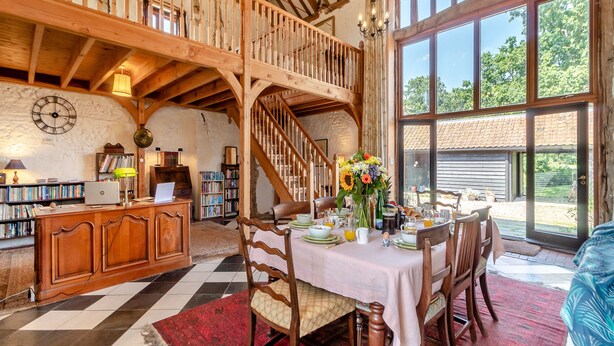 Un intérieur rustique avec un escalier en bois, un coin repas où une table est dressée pour le petit-déjeuner, et une bibliothèque remplie de livres.