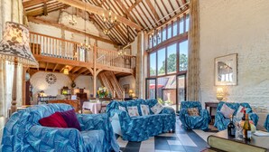 Un vaste espace de vie doté d’un plafond haut, de poutres en bois et de grandes fenêtres. On y trouve plusieurs canapés recouverts de tissus à motifs, une table à manger avec des chaises et une bibliothèque.