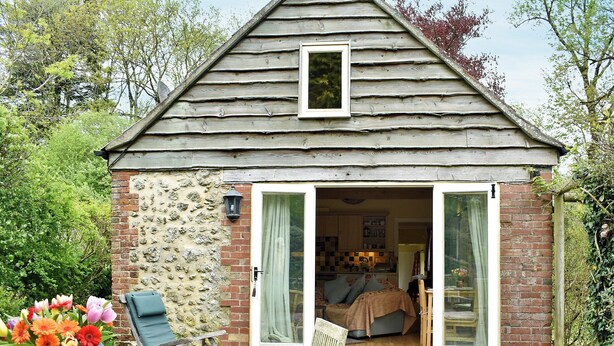 Une petite maison de campagne au charme rustique, avec une façade en pierre, un toit en pente recouvert de bois et un intérieur chaleureux comprenant un lit, un coin repas et une cuisine visible à travers une porte ouverte.