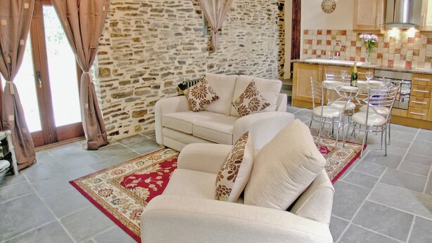 A living room with a stone wall, a sofa, a dining table with chairs, and a kitchen area with wooden cabinets. - Lower Shippen - 23493 (North Petherwin, near Launceston)