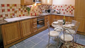 A kitchen with wooden cabinets, a stainless steel range hood, a microwave, and a sink. There is a dining table with chairs and a vase with flowers. - Lower Shippen - 23493 (North Petherwin, near Launceston)
