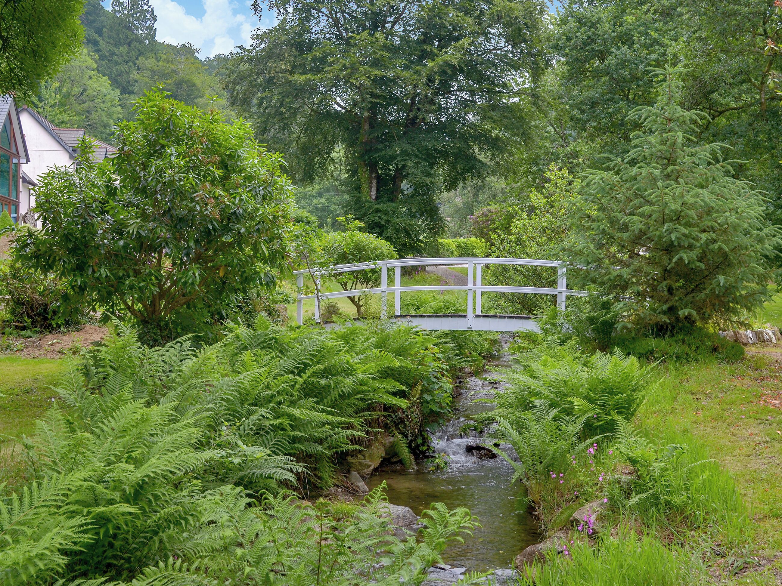 Een witte brug over een beek in een weelderige tuin met dichte begroeiing en bomen.
