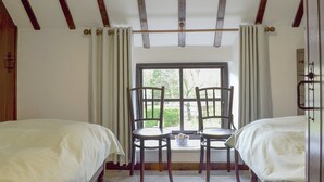 A room with two beds, a window with curtains, a chair, and a small table with a bowl.