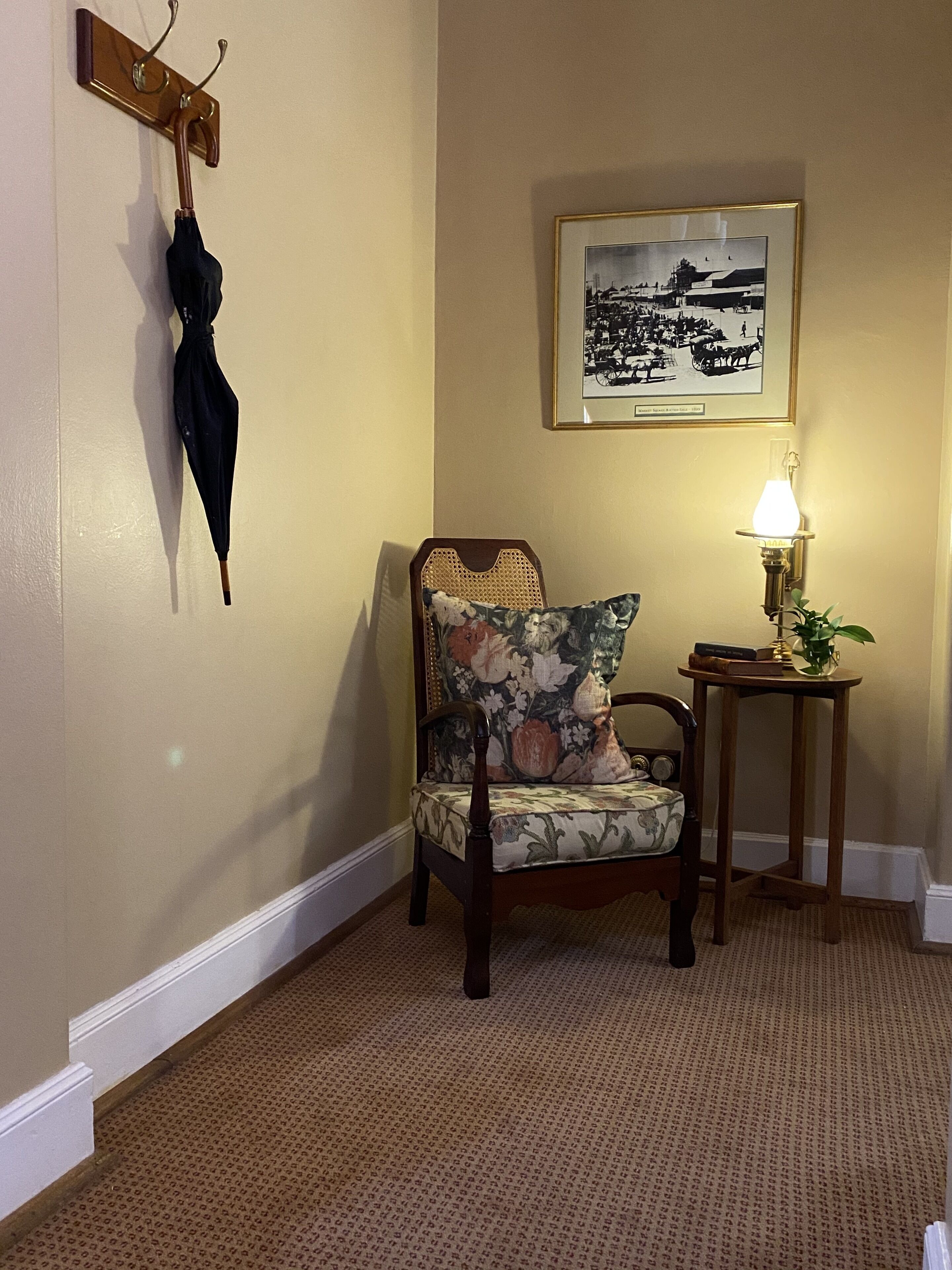 A corner room with a wooden chair, a small table with a lamp, a framed picture on the wall, and a coat rack with an umbrella.