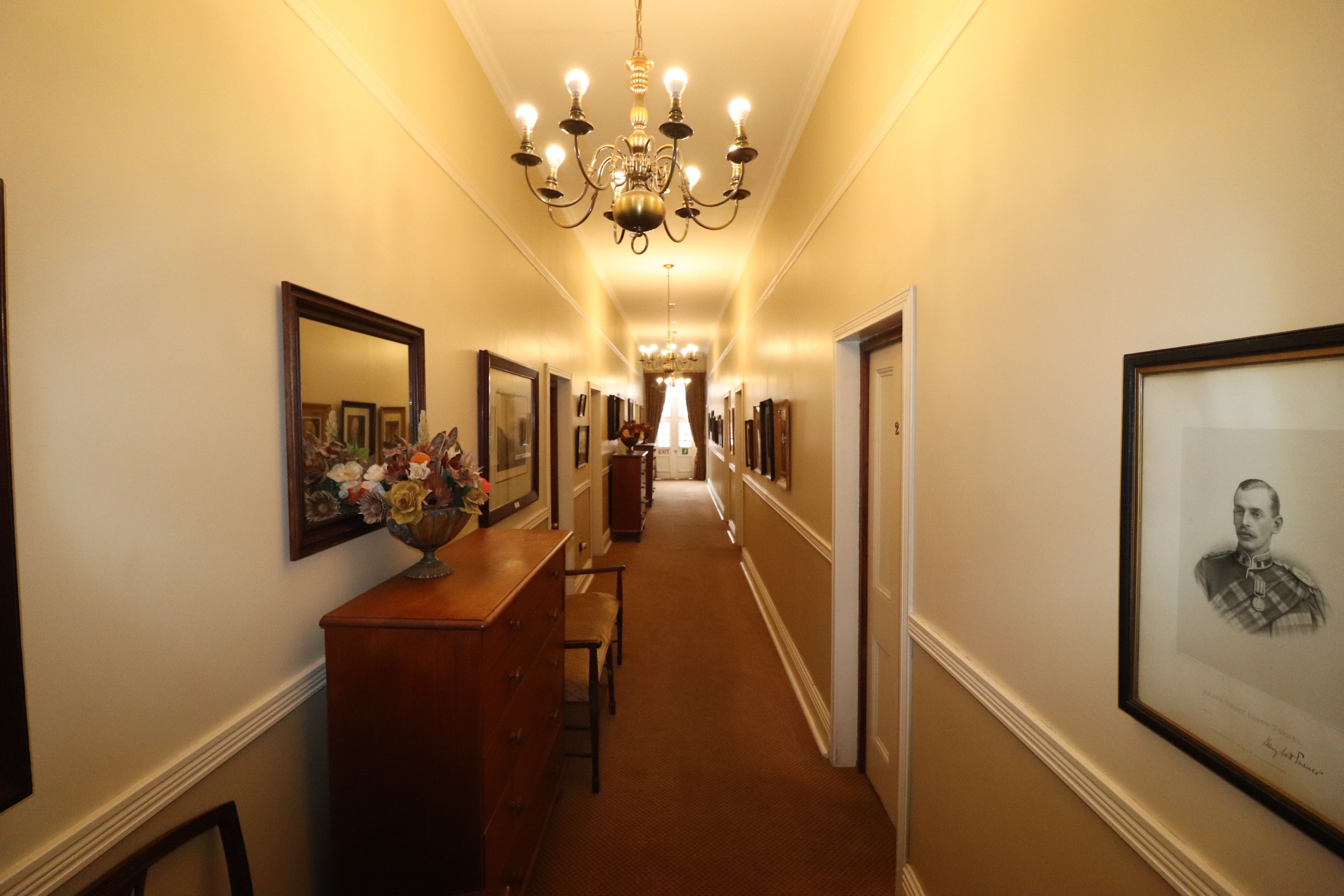 A hallway with a chandelier, framed pictures, and a wooden dresser with a vase of flowers.