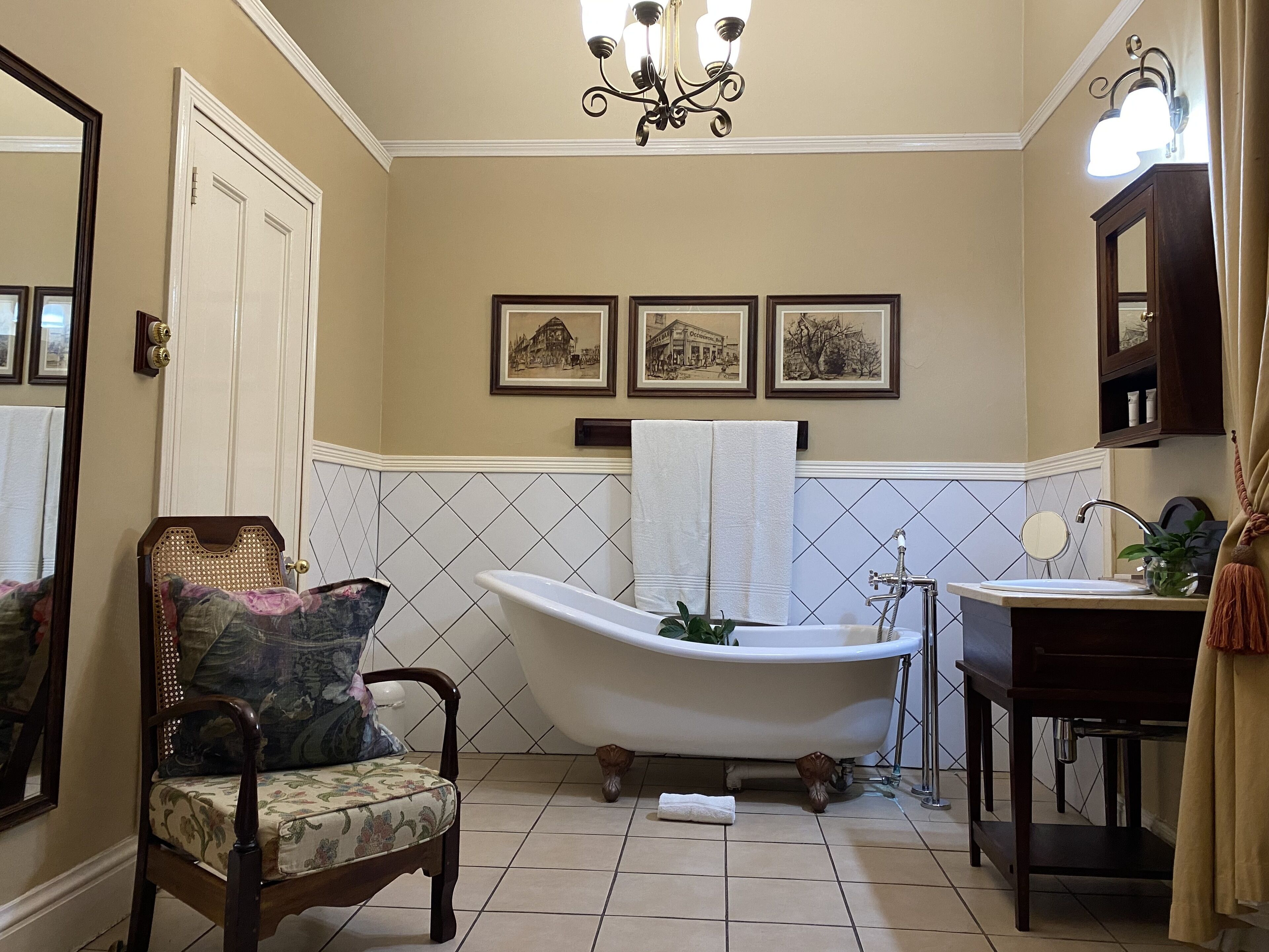 A bathroom with a clawfoot bathtub, a wooden chair with a floral cushion, a sink with a mirror, and framed pictures on the wall.
