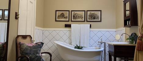 A bathroom with a clawfoot bathtub, a wooden chair with a floral cushion, a sink with a mirror, and framed pictures on the wall.