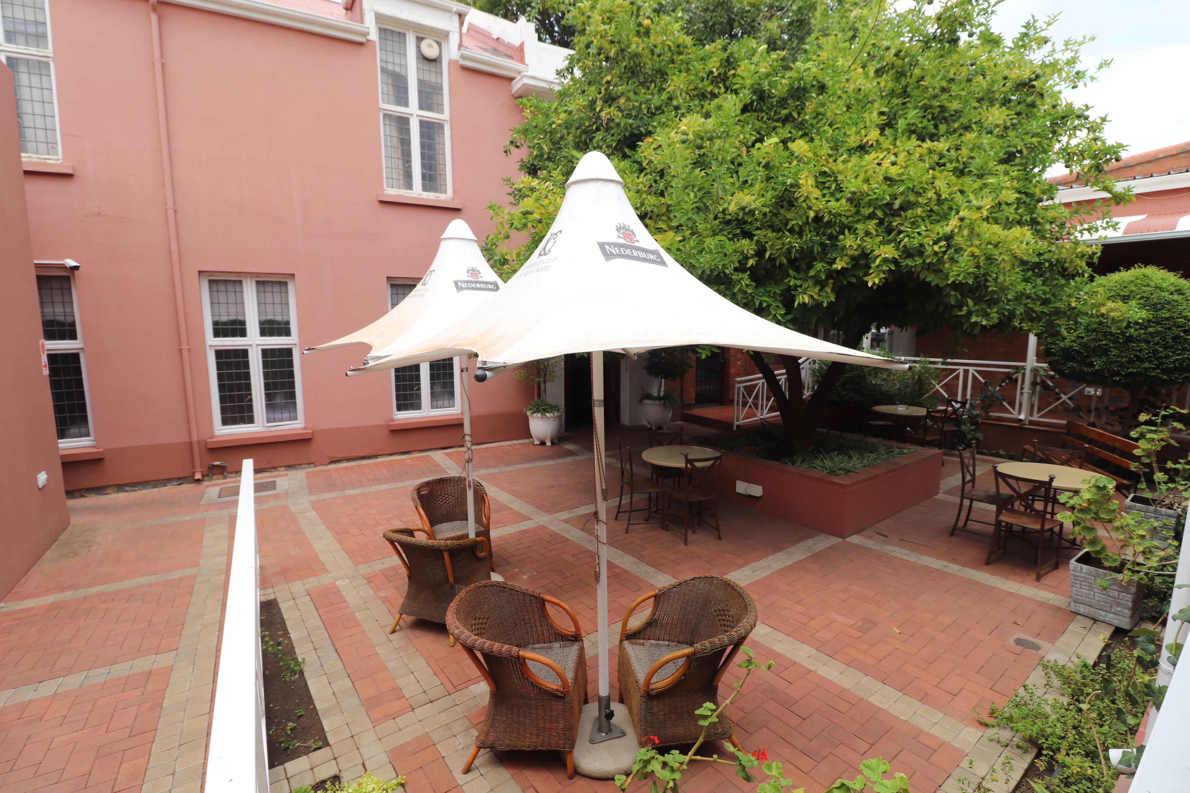 A courtyard with wicker chairs and tables under white umbrellas, a brick floor, and a building with a pink facade.
