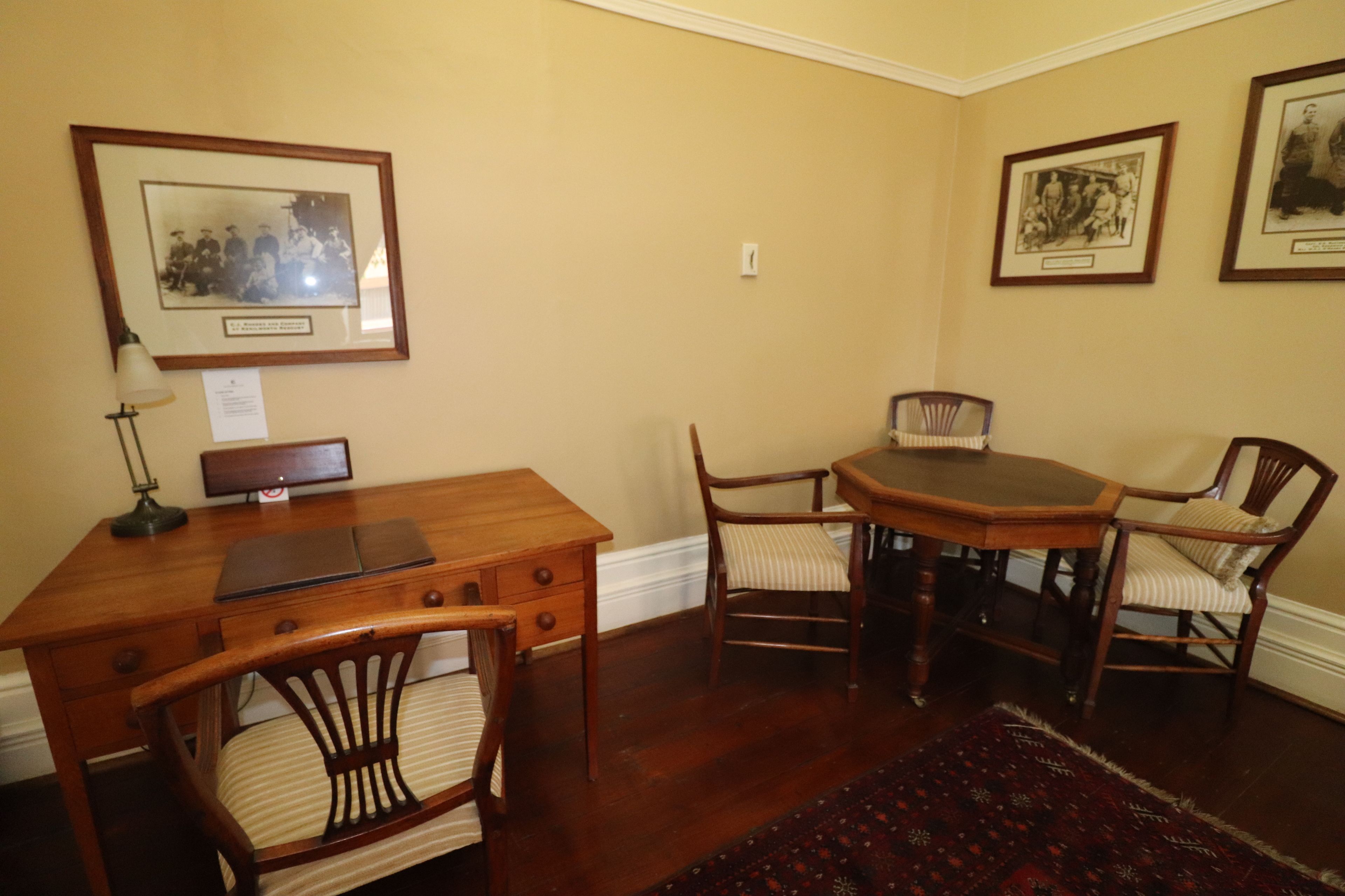 A dining area with a wooden table and chairs, framed pictures on the walls, and a desk with a lamp.