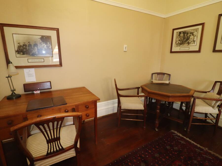 A dining area with a wooden table and chairs, framed pictures on the walls, and a desk with a lamp.