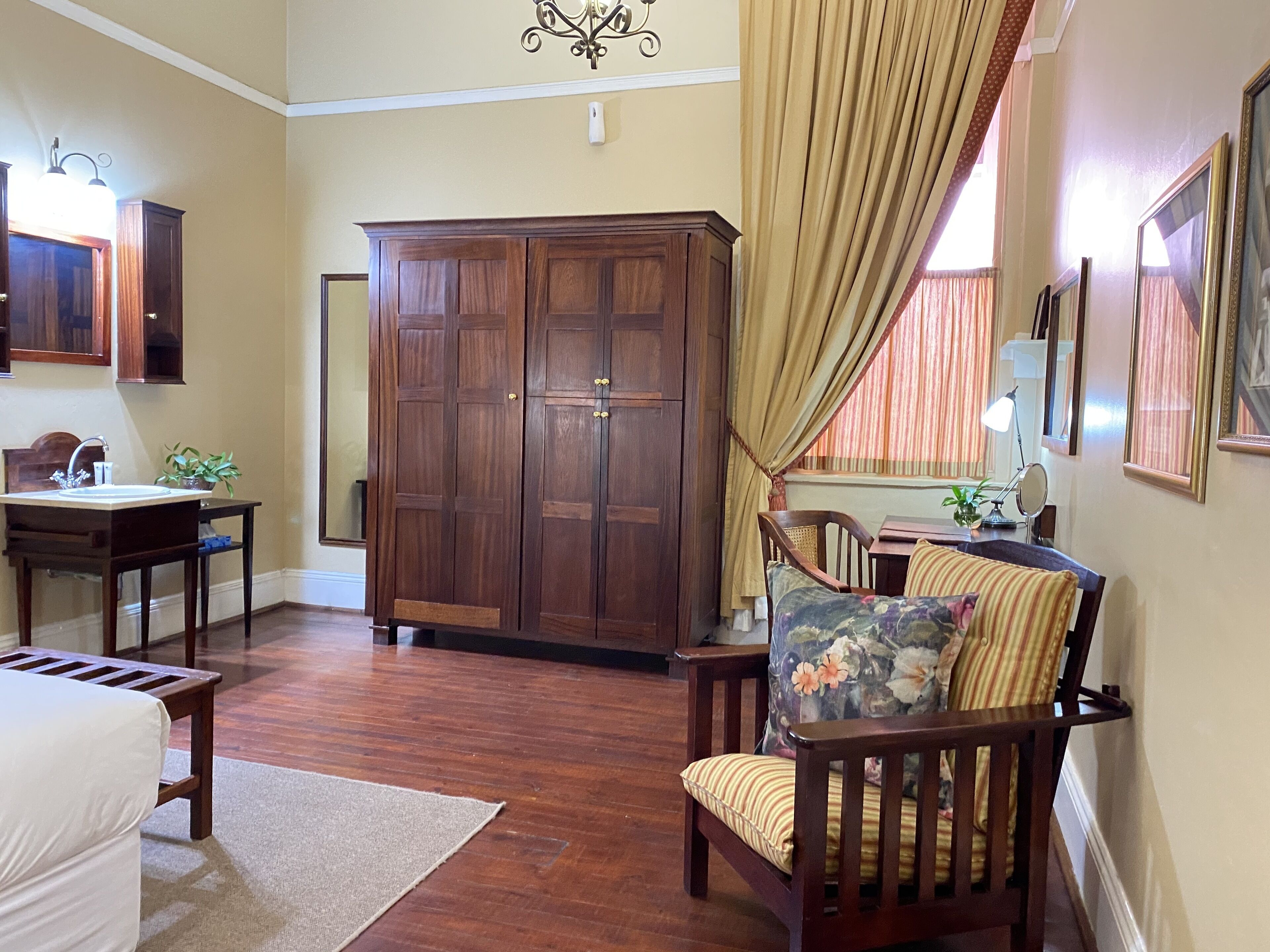A room with a wooden wardrobe, a desk with a chair, a bed with a white bedspread, and a large mirror on the wall.