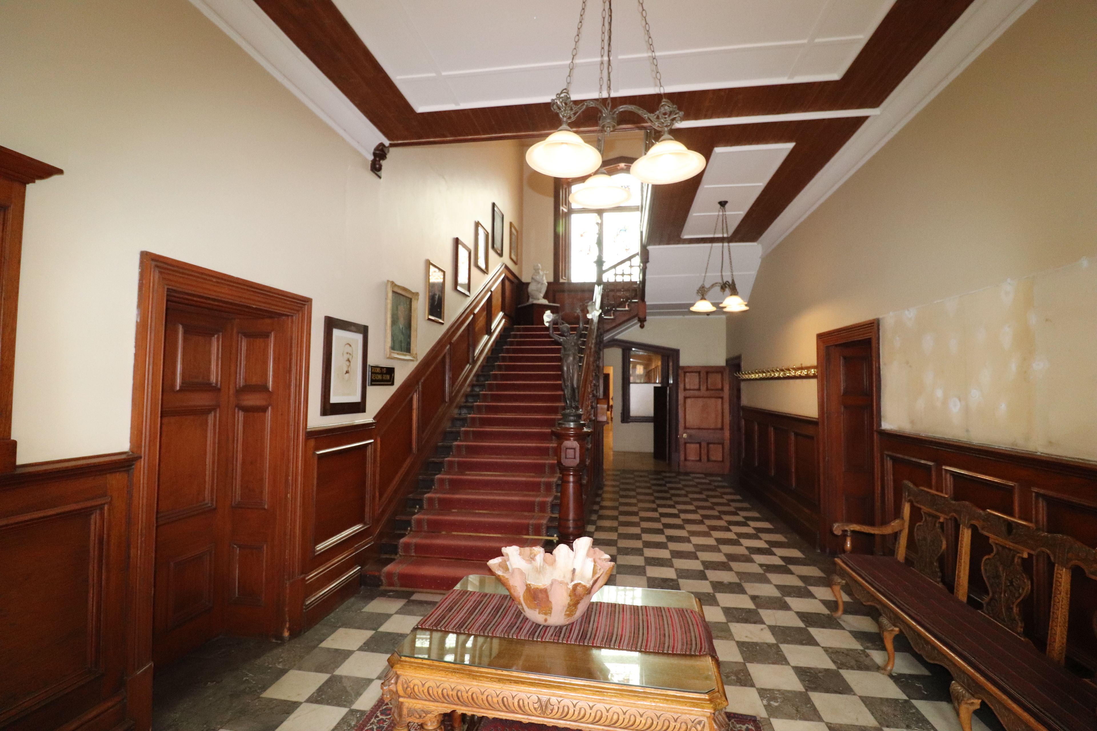 A grand hallway with a wooden staircase, a checkered floor, and a chandelier.