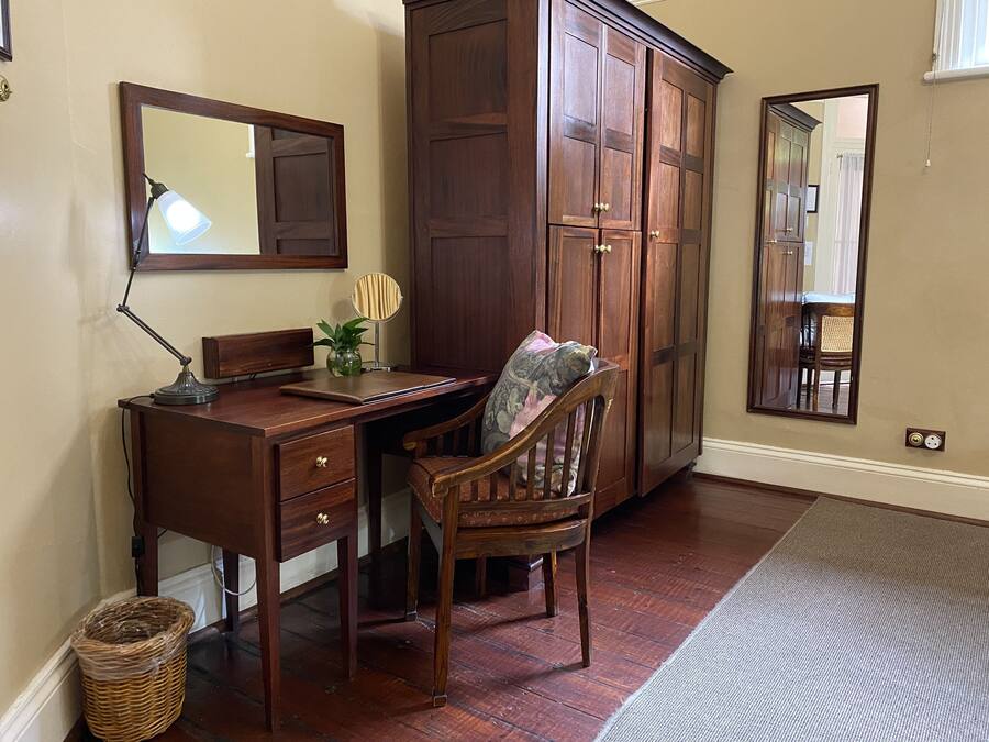 A wooden desk with a lamp, a chair, a mirror, and a cabinet in a room with wooden flooring and a rug.