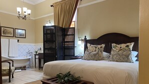 Une chambre à coucher avec un grand lit, une table de chevet en bois, une petite table avec une plante, une baignoire de style classique et une tête de lit en bois foncé.