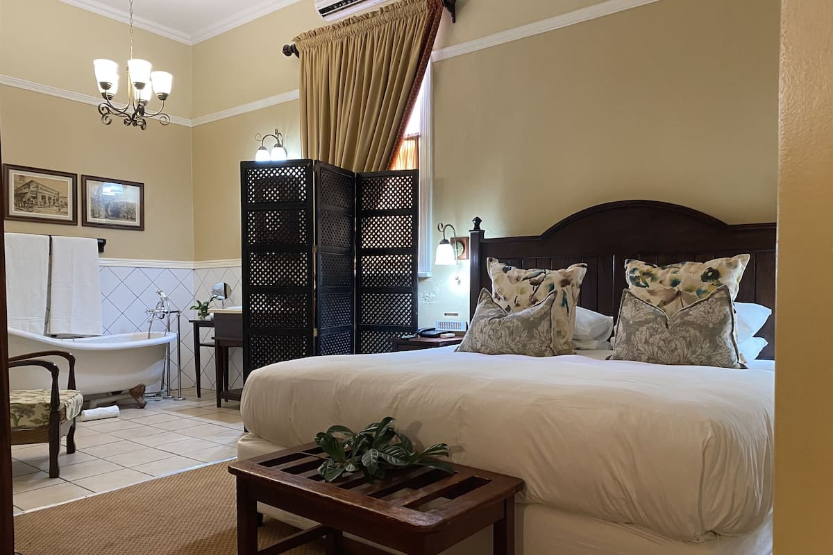 Une chambre à coucher avec un grand lit, une table de chevet en bois, une petite table avec une plante, une baignoire de style classique et une tête de lit en bois foncé.