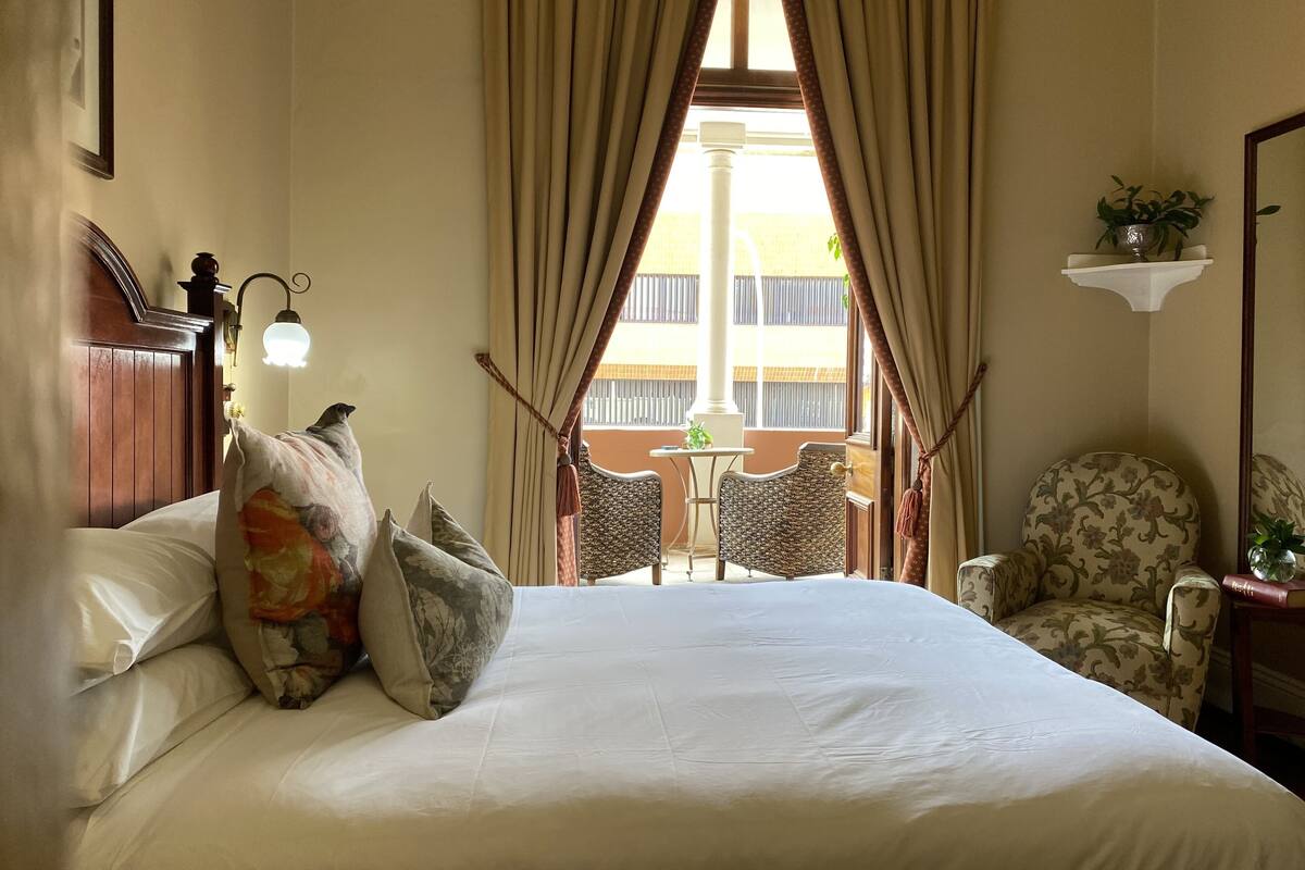Une chambre d’hôtel avec un grand lit, une chaise, une petite table et une fenêtre avec des rideaux.
