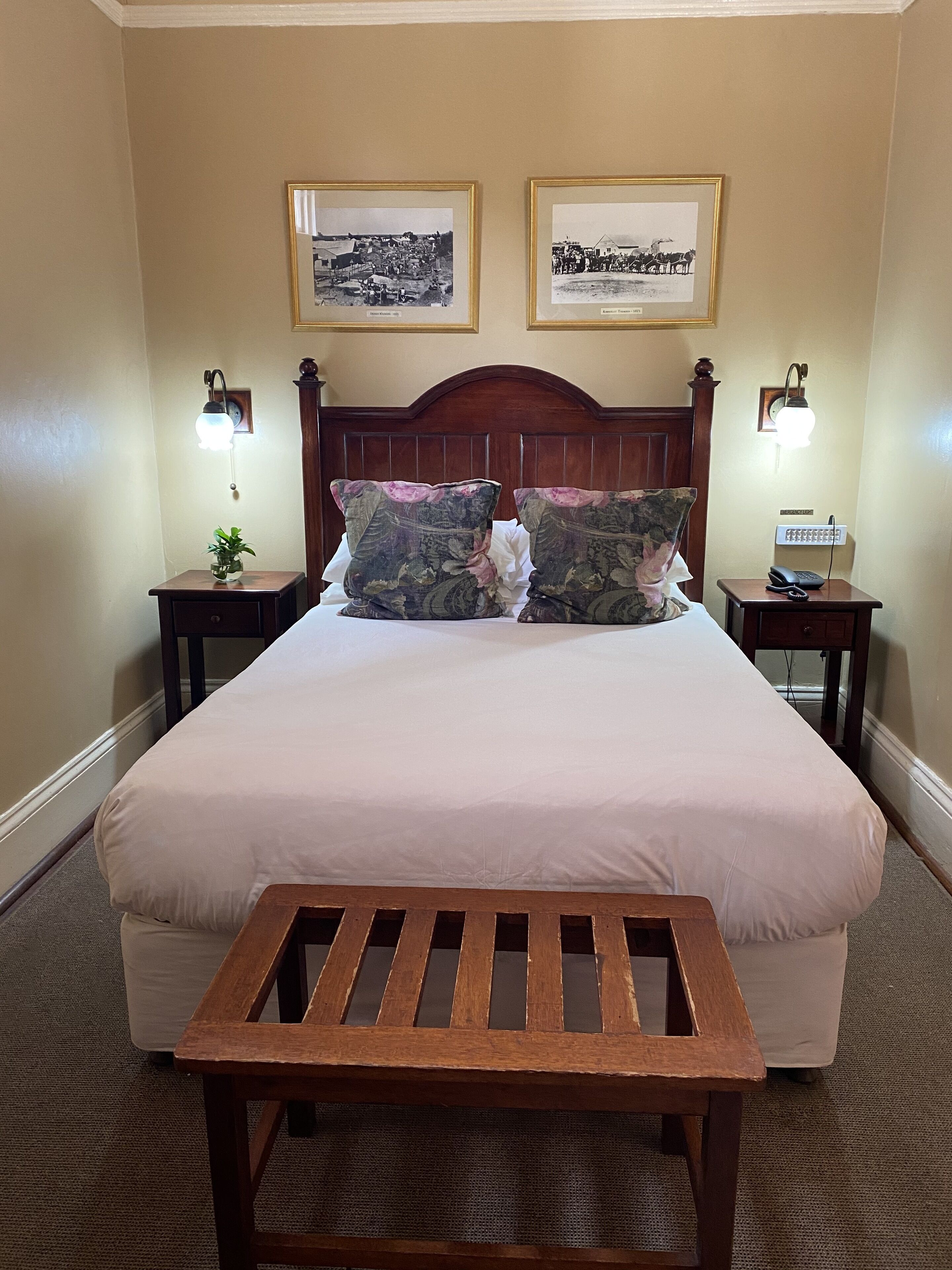 Habitación de hotel con una cama grande, dos mesitas de noche, un banco de madera y dos cuadros en la pared.