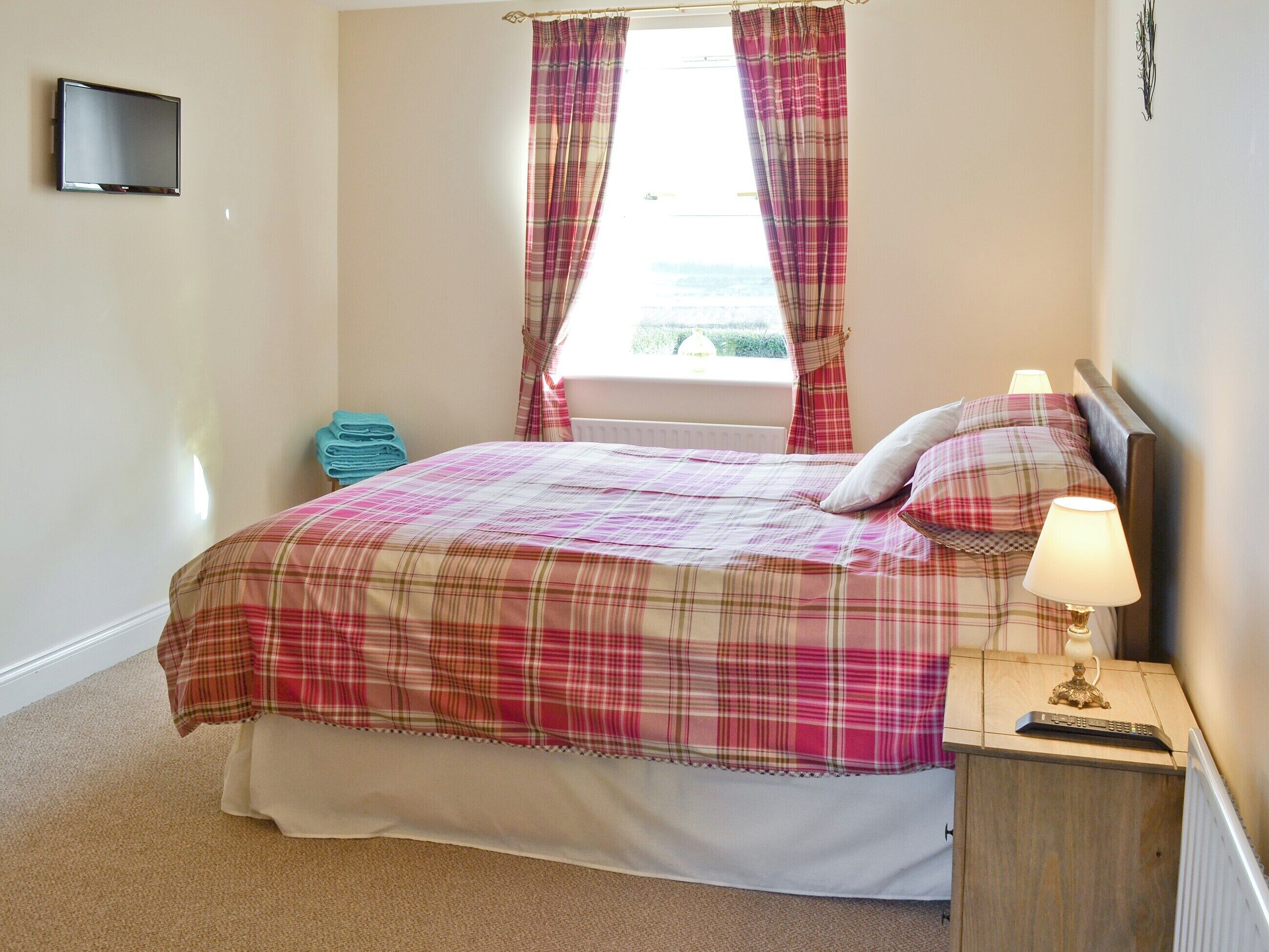 A bedroom with a bed, a nightstand with a lamp, a TV on the wall, and a window with curtains.