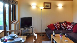 Una sala de estar acogedora con un televisor sobre un pedestal de vidrio, un sofá con almohadas rojas y blancas, una mesa de comedor con platos y una botella de vino, y una lámpara de pie.