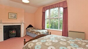 A bedroom with a fireplace, a bed with floral bedding, a window with curtains, and a hanging light fixture. - 3 bedroom accommodation in Waxham (Waxham)
