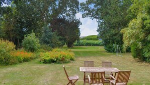 An outdoor seating area with a wooden table and chairs, surrounded by lush greenery and a hedge. - 3 bedroom accommodation in Waxham (Waxham)