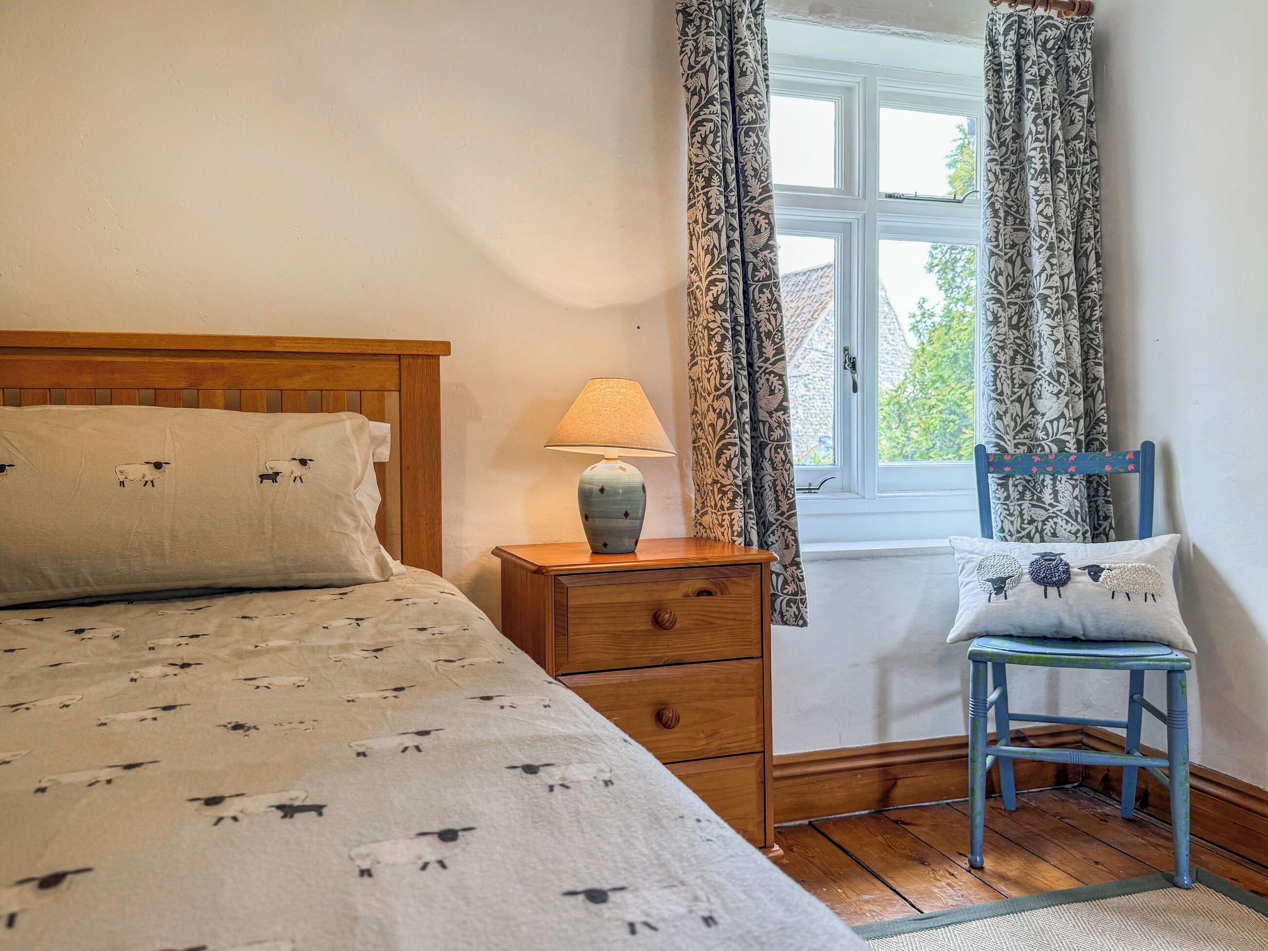 Een slaapkamer met een bed, nachtkastje, lamp, stoel en een raam met gordijnen.