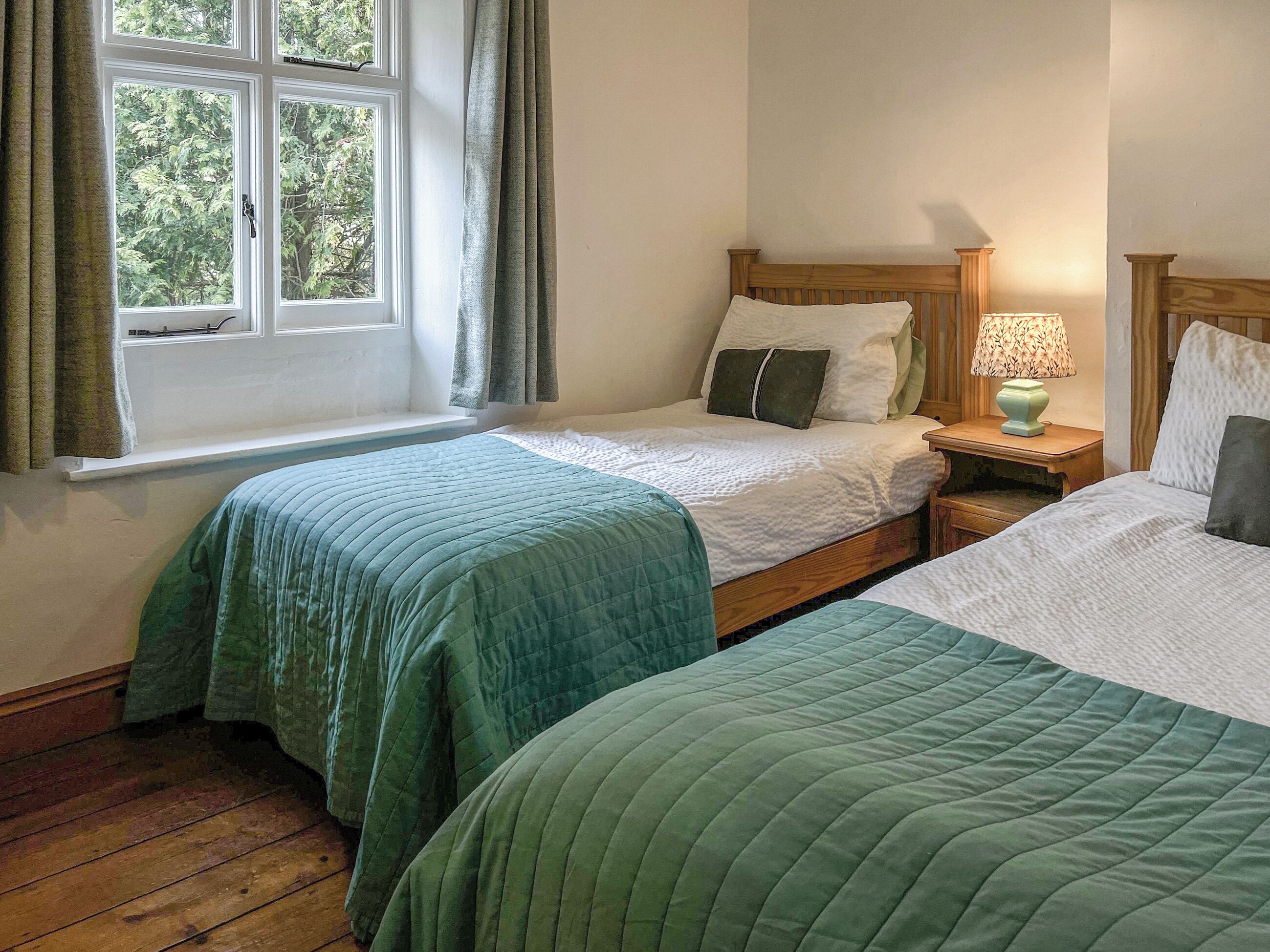 A room with two single beds, a window with curtains, a bedside lamp, and a view of trees outside.