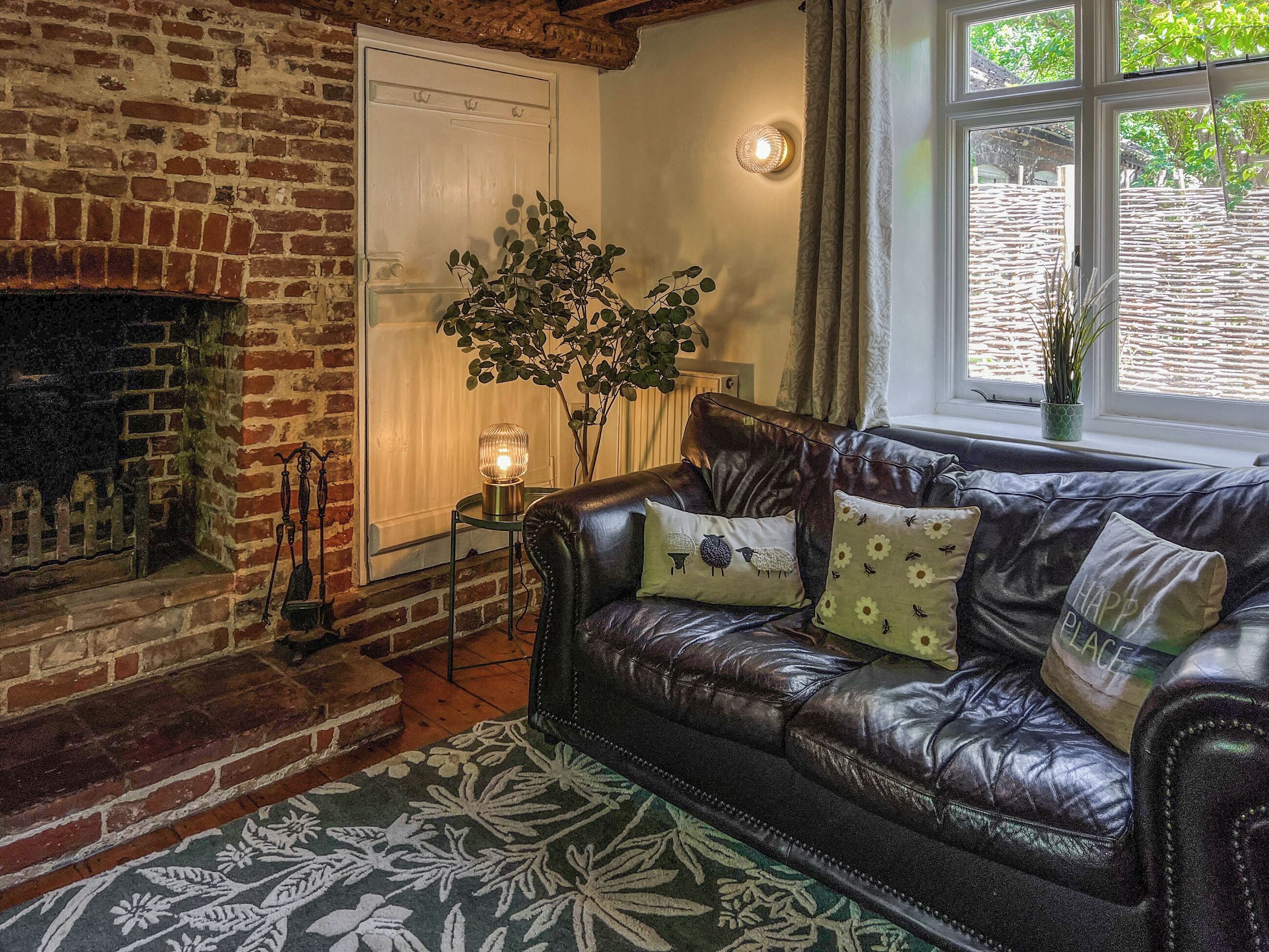 A cozy living room with a leather sofa, a fireplace, a potted plant, and a view of greenery through a window.